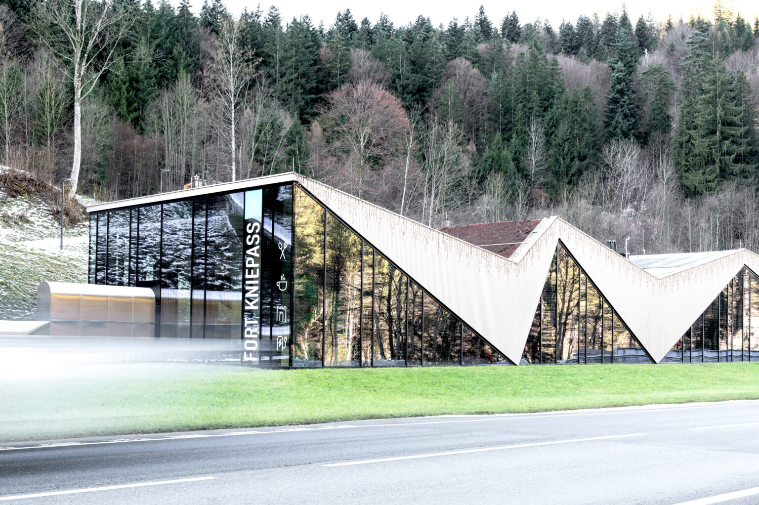 Moderne Glasfassade des Besucherzentrums Fort Kniepass in Unken mit markanter Zick-Zack-Dachkonstruktion und Stahl-Pfosten-Riegel-Elementen vor Waldhintergrund.